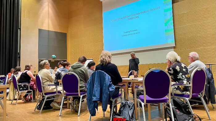 Bilde av en forsamling med folk i en konferansesal foran en storskjerm