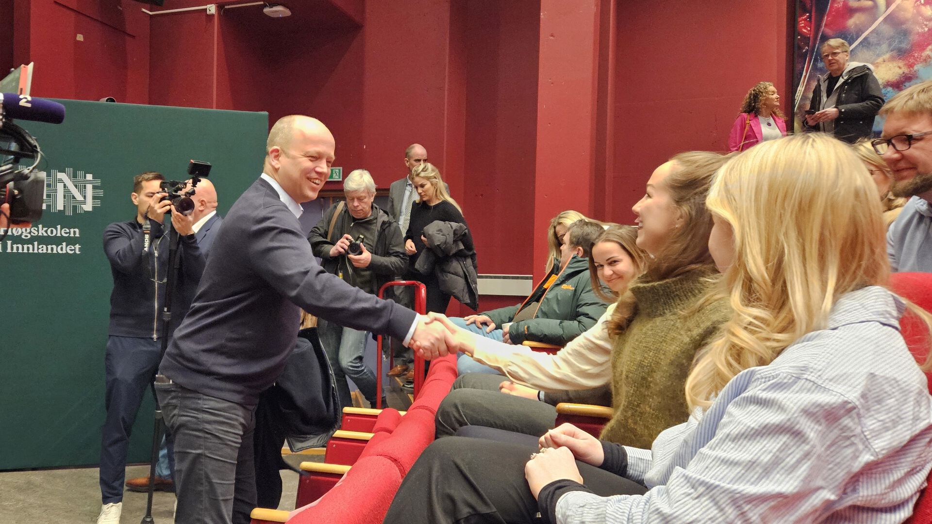 Finansministeren smiler og håndhilser på studenter på første rad, fotografer tar bilder.