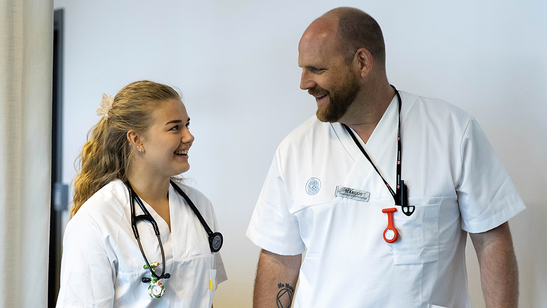To sykepleierstudenter en mann og en dame ser på hverandre og smiler. Begge har på hvit sykepleier uniform.