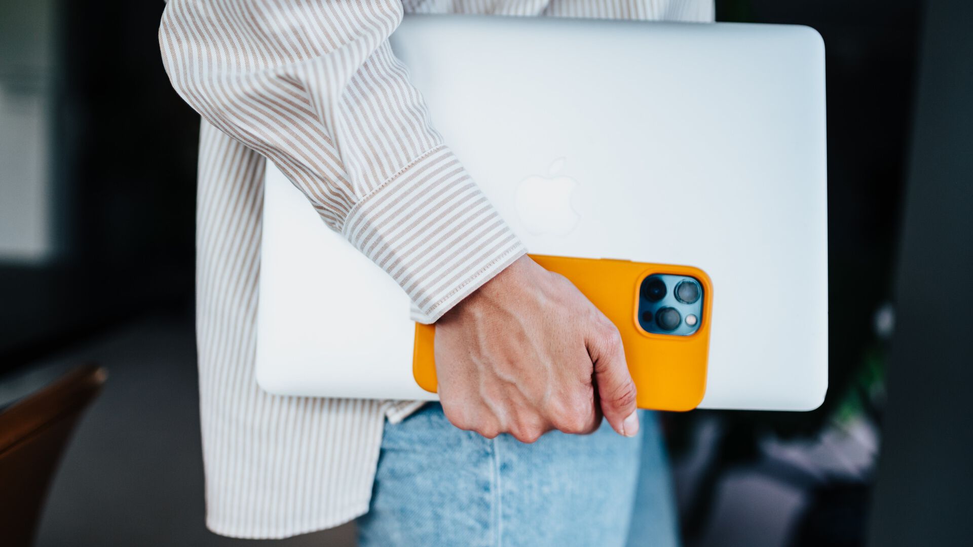 The arm and hand of a person in jeans and a shirt is holding a laptop and a phone in a yellow case close to them while walking.