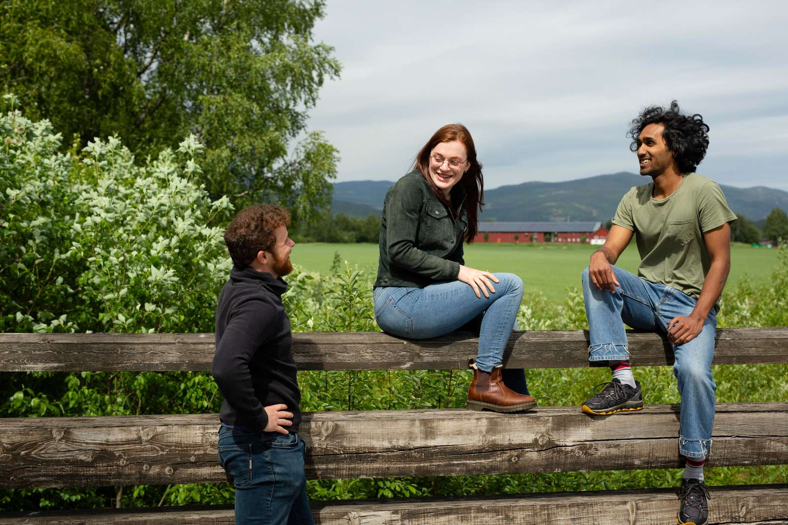 To personer, en jente og en gutt sitter på et gjerde, en annen gutt står ved siden av og ser opp på de. De smiler til hverandre. Det er grønt og frodig rundt de og en rød låve skymtes i bakgrunnen.