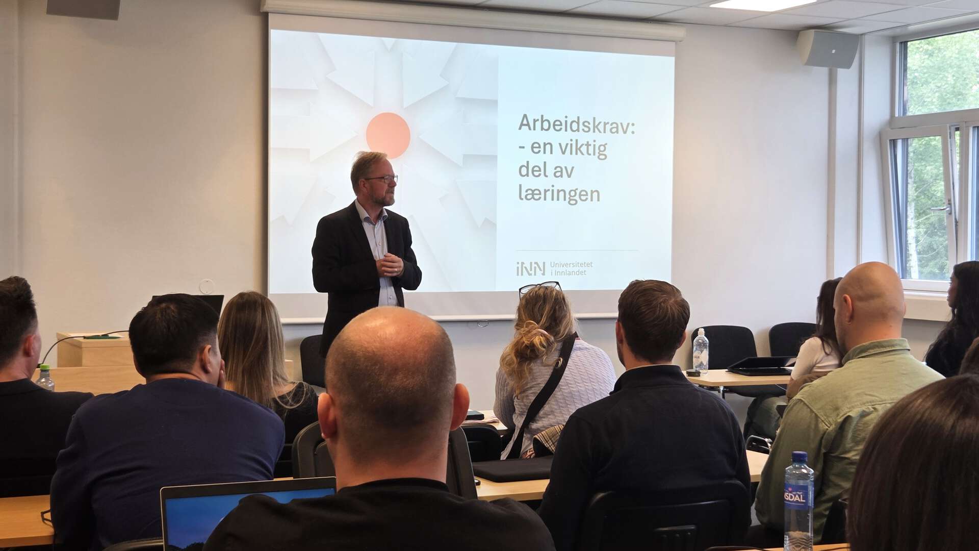 Professor Bård Tronvoll foreleser, mange studentrygger i forgrunnen.Tekst på lysbildepresentasjonen er Arbeidskrav - en viktig del av læringen