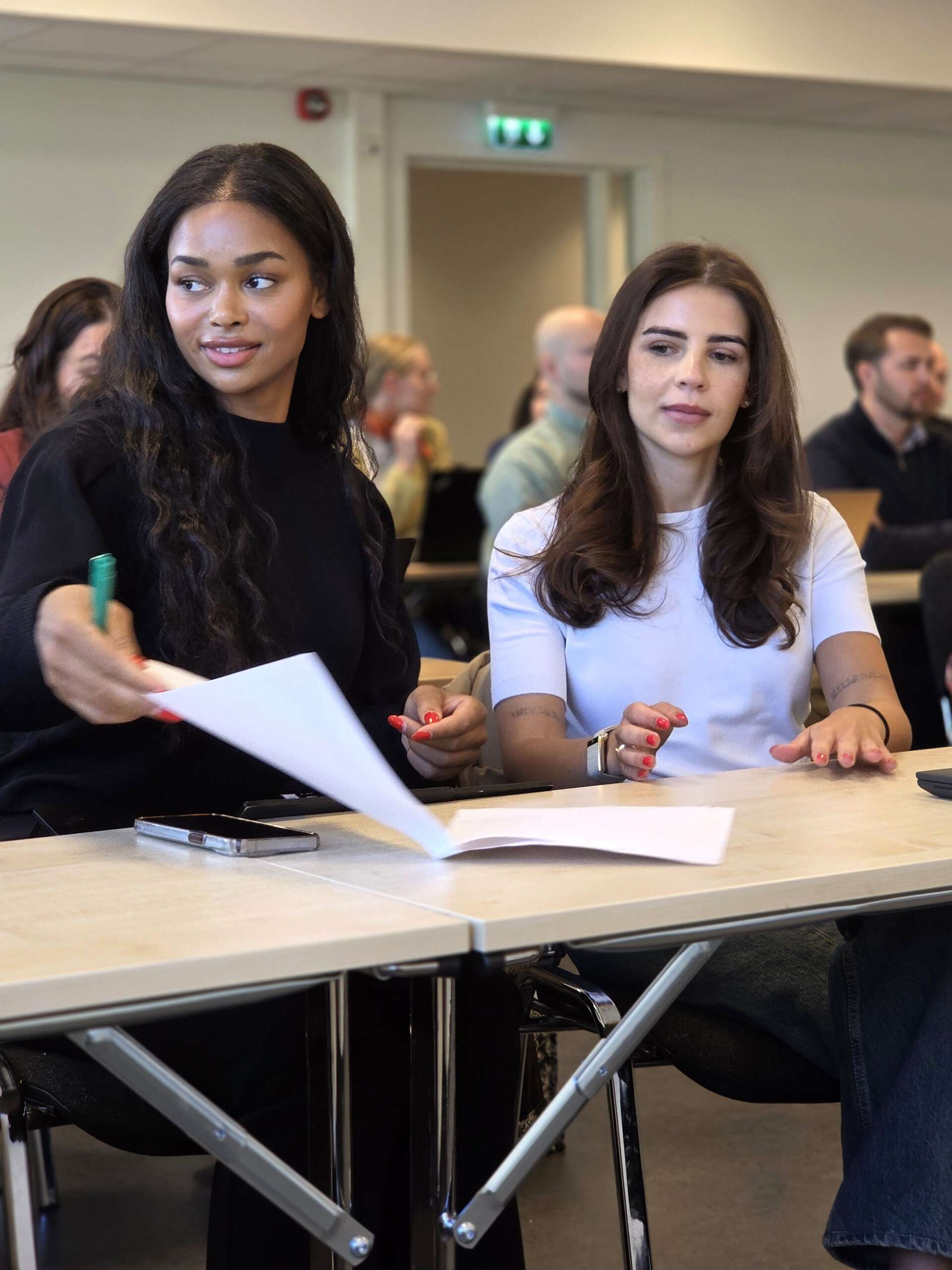 To studenter som sitter ved siden av hverandre og blar i papirer på pulten.