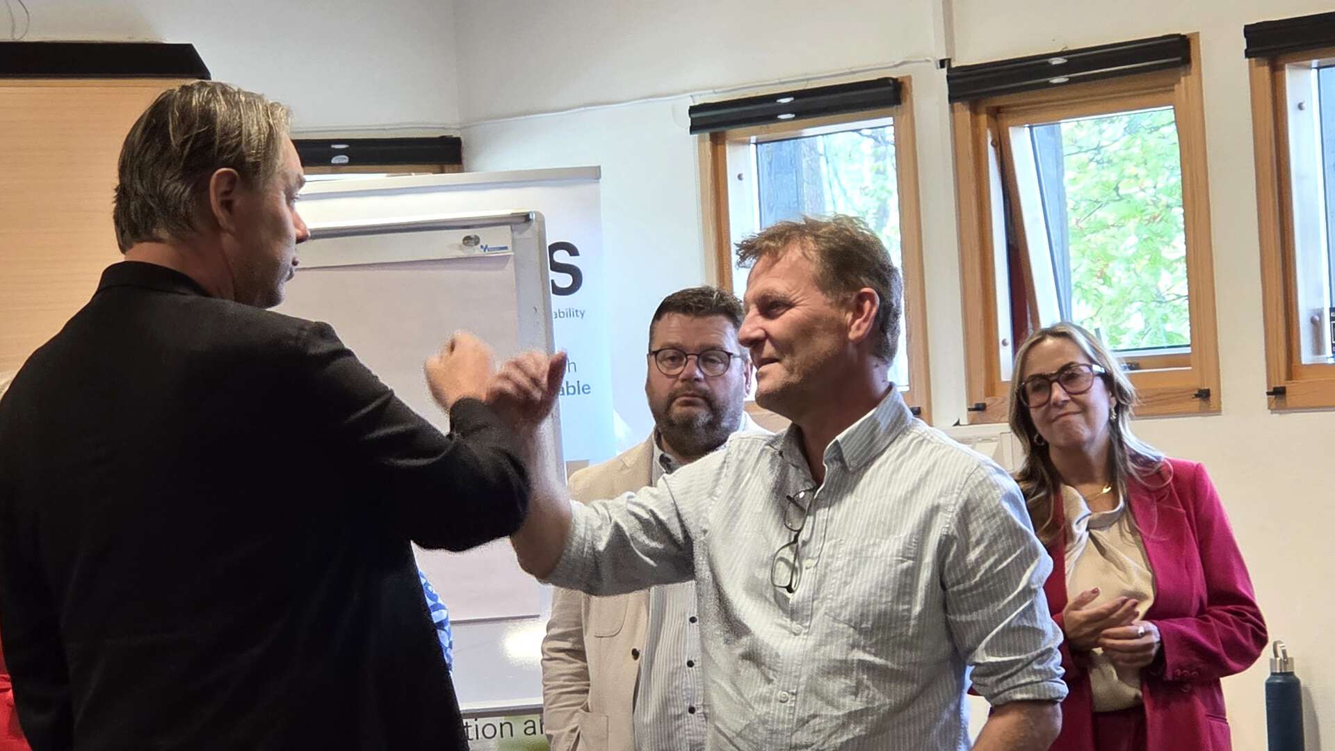 To menn hilser med en håndhilsen i et møterom, mens en mann og en kvinne ser på i bakgrunnen. Nakne vinduer og en flippover står langs veggen.