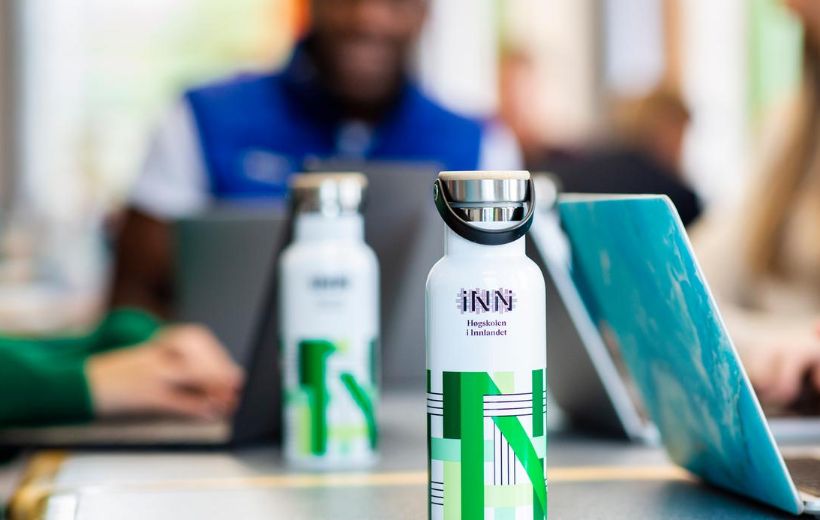 Flasker med HINN-logo i forkant, mennesker med pc uklart i bakgrunnen.