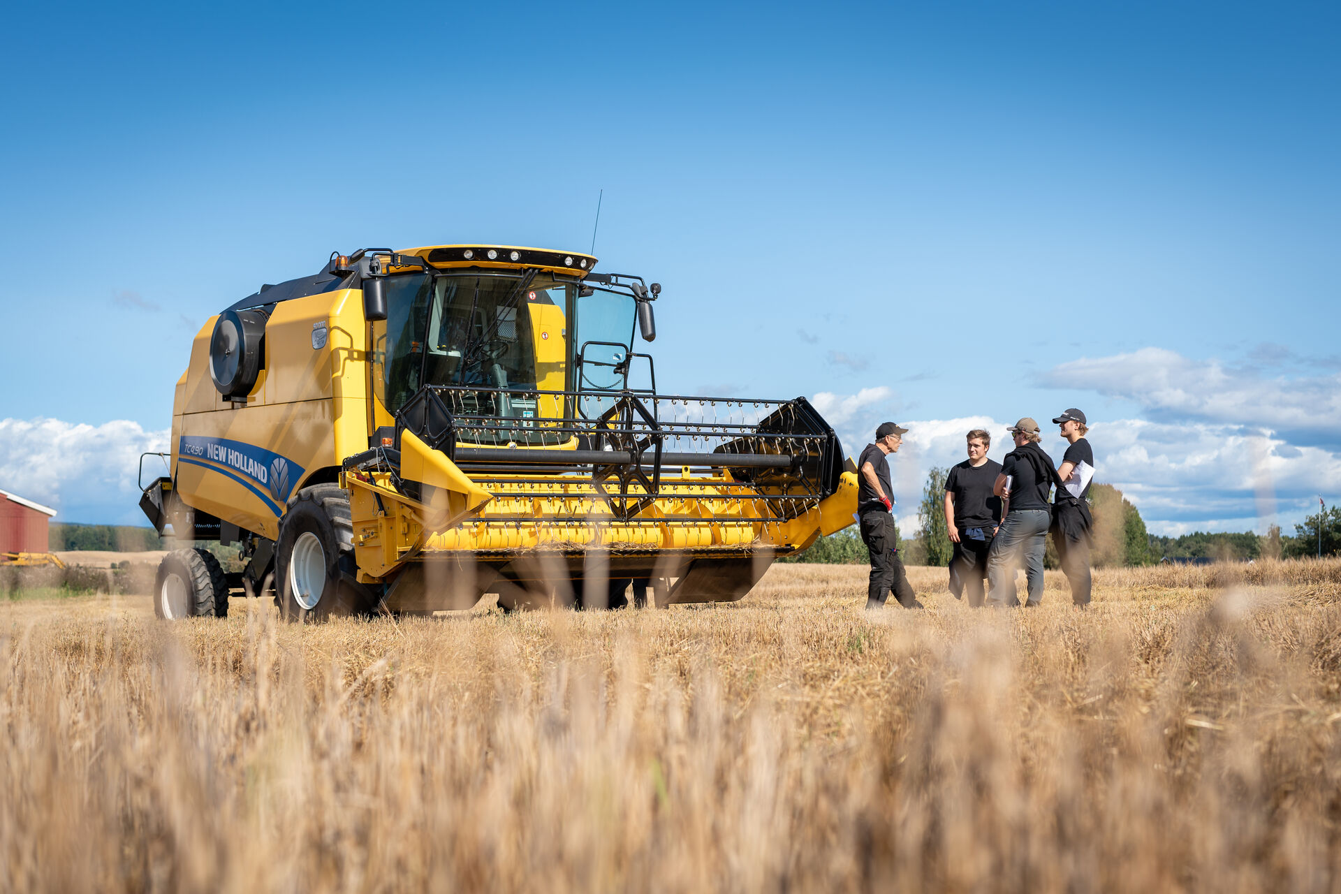 An agricultural machine stands in the cornfield at Blæstad. Four students are standing next to it, talking to each other.
