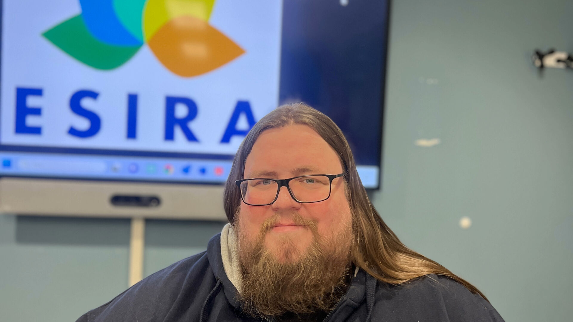man with long hair and beard, smiling in front of colourful logo wih abbreviation ESIRA