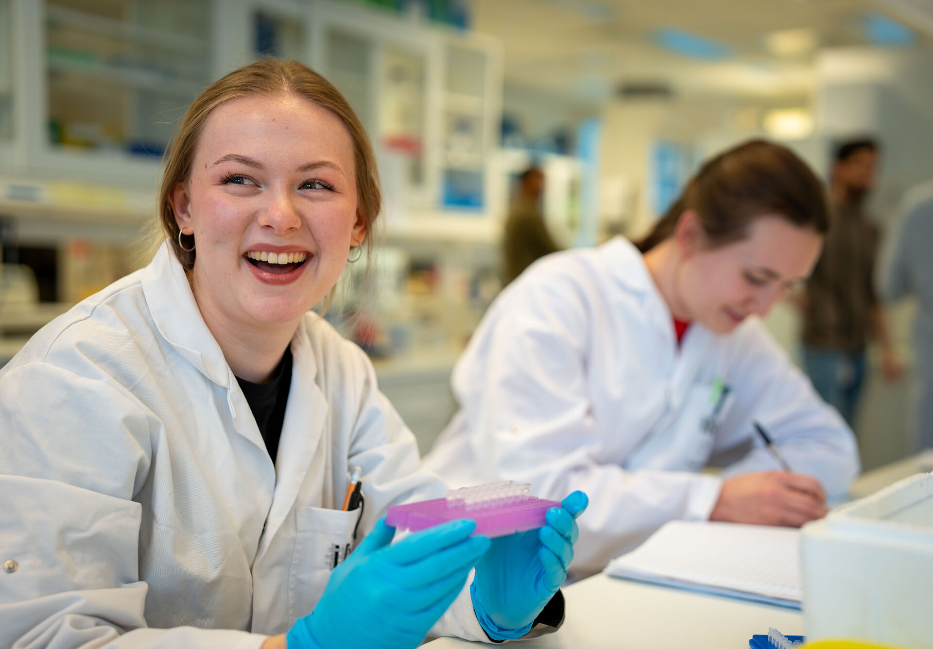 To kvinnelige studenter sitter i et laboratorium, iført hvite labfrakker. Hun nærmeste smiler og holder opp et brett med små pipetter, brettet er lilla og hun har på seg blå hansker. Hun bakerste er ikke i fokus, men sitter og skriver på noe.