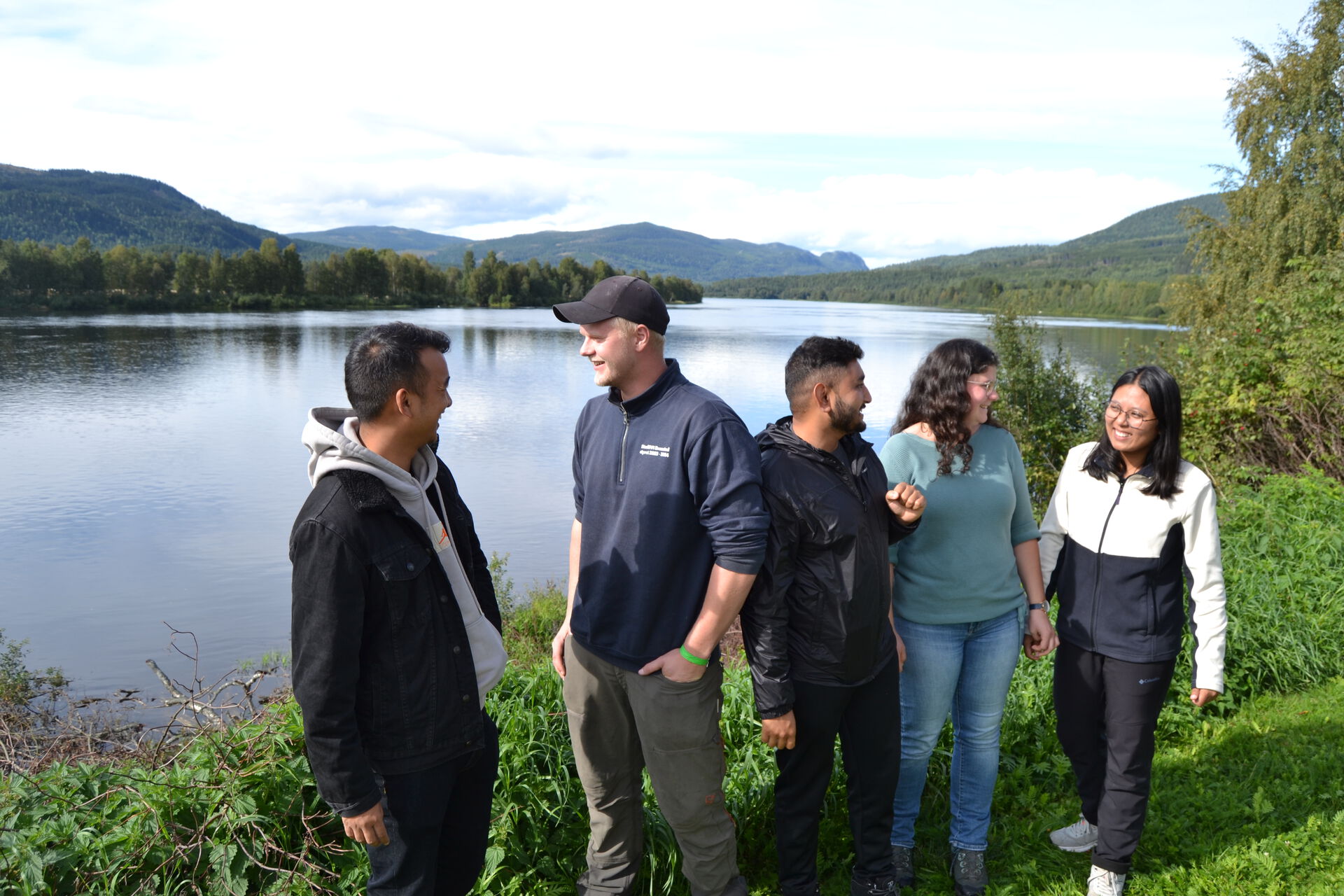 Five students are talking and smiling. They are standing by a river with green plants around them