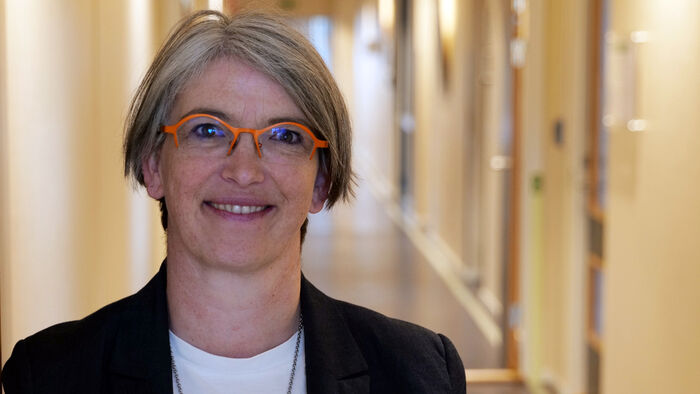 A woman with short blond hair and red-rimmed glasses standing in a corridor smiling.