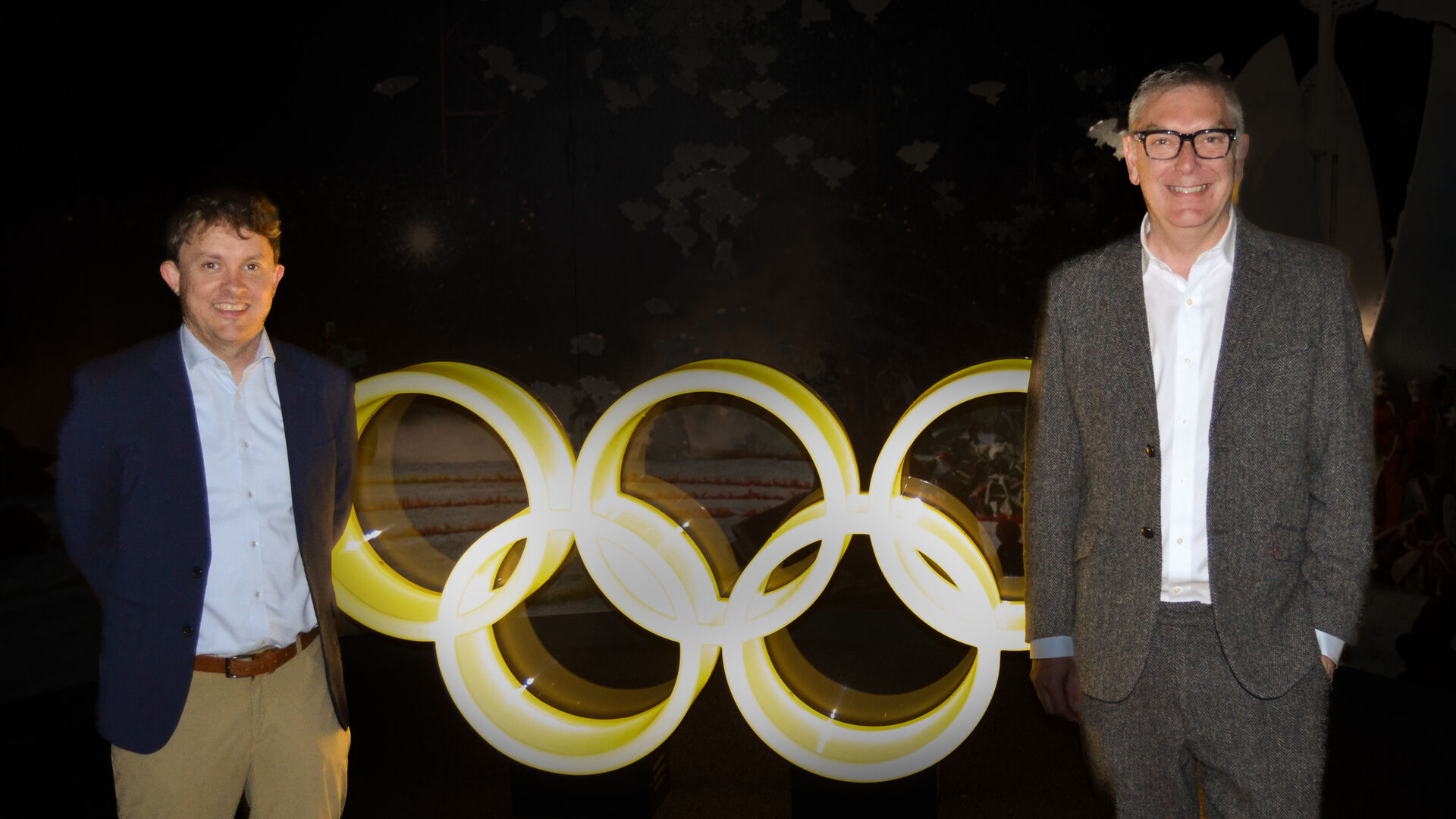 Jörg and Mark are standing in front of the bright Olympic rings in a dark room