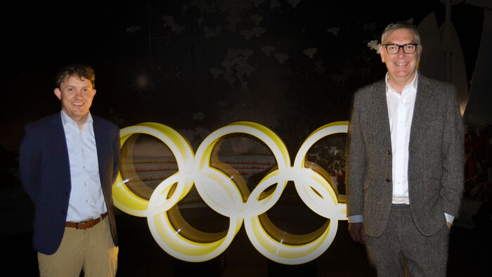 Jörg and Mark are standing in front of the bright Olympic rings in a dark room