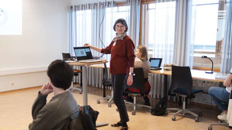 A woman is standing in front of an open laptop, with three other people in the background