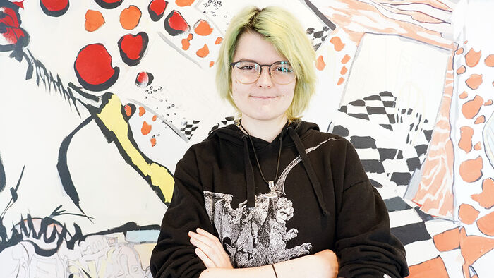 Portrait photo of a female student dressed in black infront of a wall covered in graffiti. 