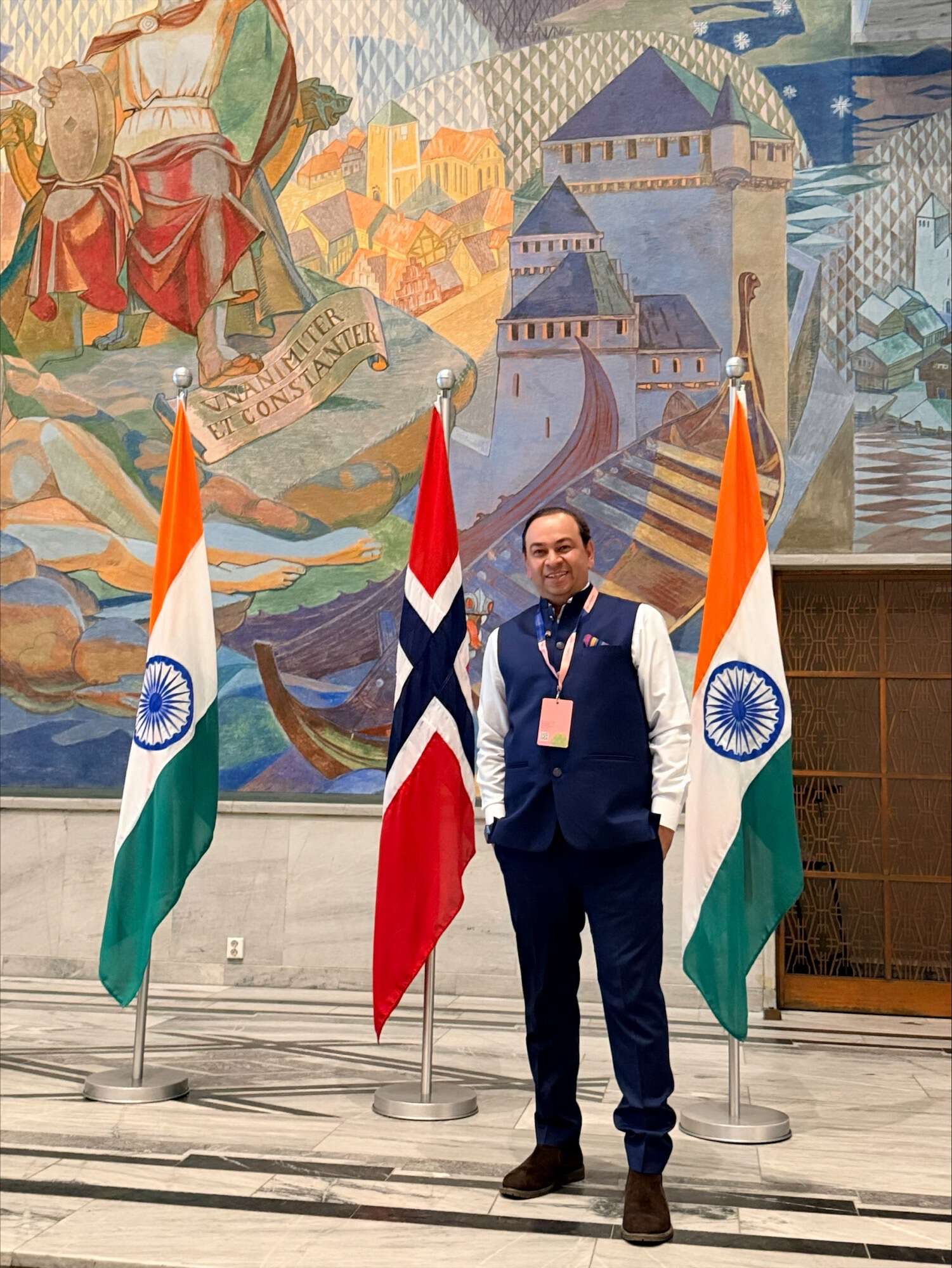 A smiling man wearing a white shirt, blue vest and black pants standing with two Indian flags and one Norwegian flag behind him with a wall mural in the background.