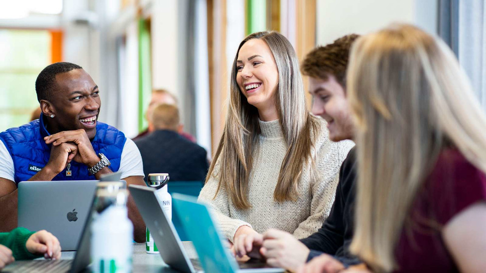 Four smiling students, each with a laptop, talking around a table. 