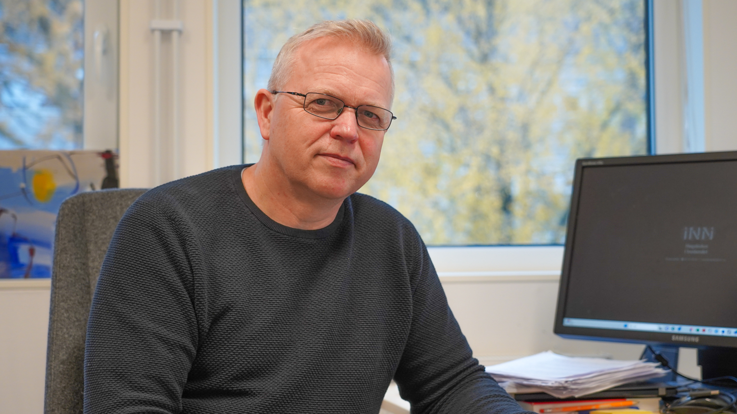 Per Kristian in front of his computer and with a window in the background where ypu can see a autumn-yellow three