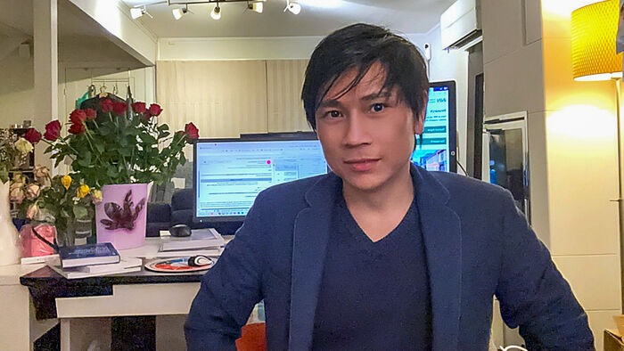 Christopher in a room with flowers and two big screnes in the background