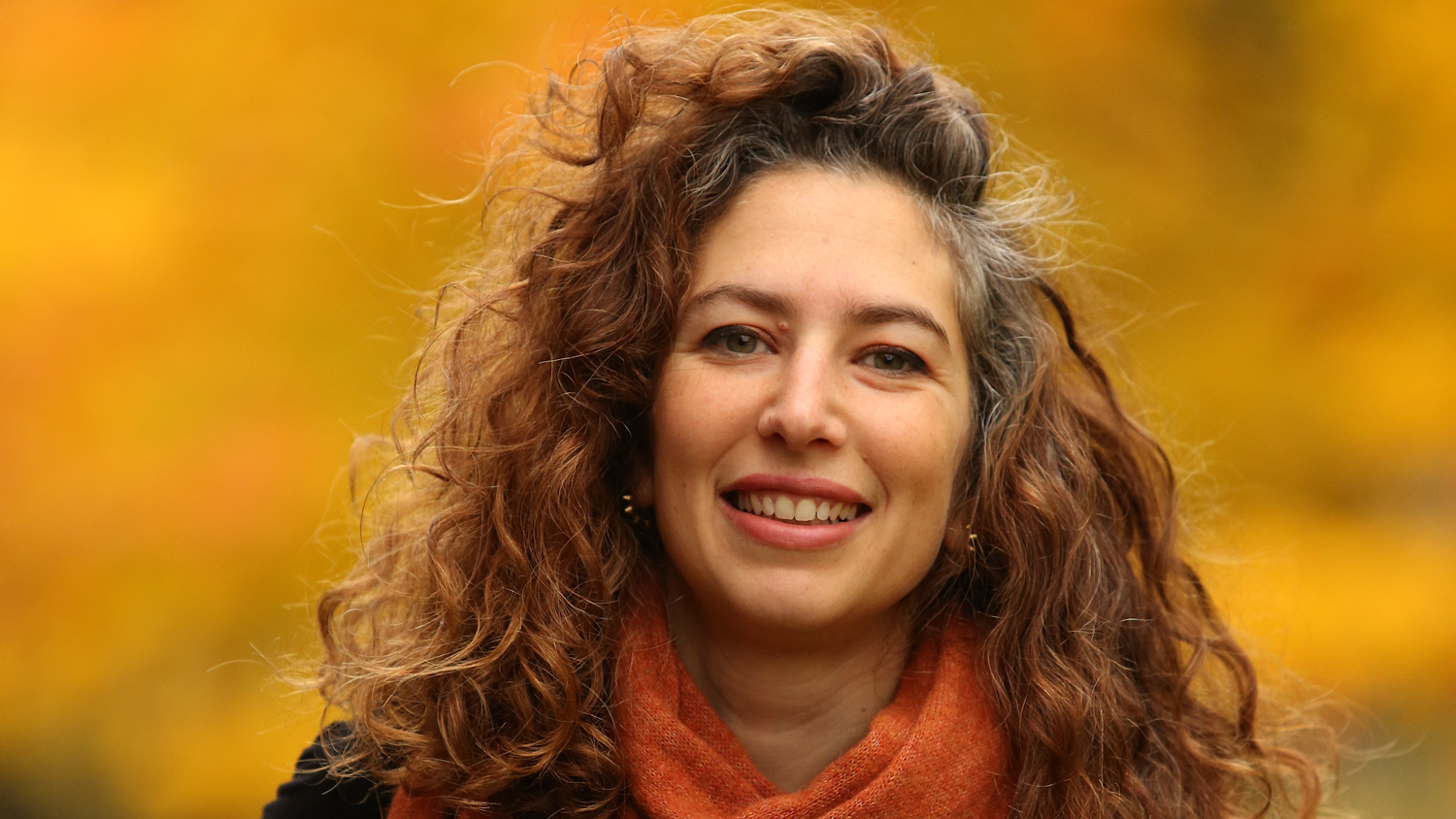 Portrait of Dalia AlKury with an orange background. She is smiling and wearing an orange scarf