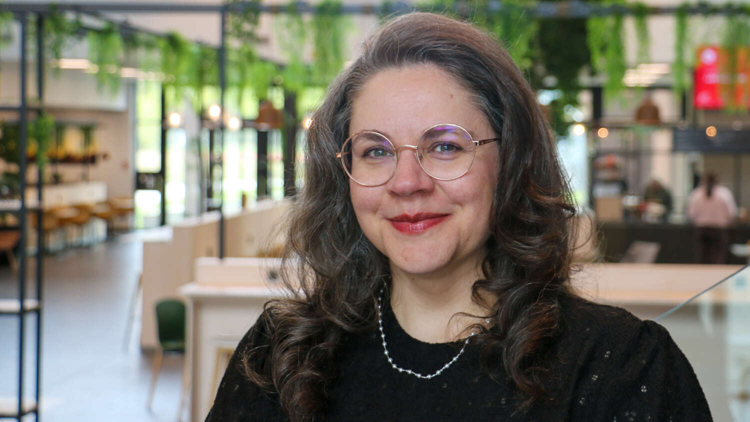 A smiling woman with long brown hair wearing glasses and a black sweater smiling at the camera with a blurred background.