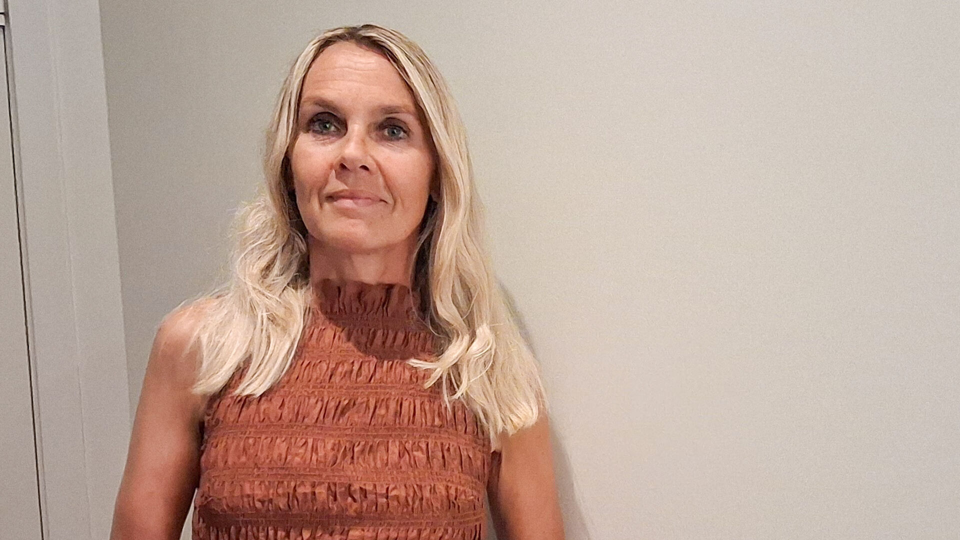 Portrait photo of a woman with blonde hair and a rust-coloured dress against a light wall