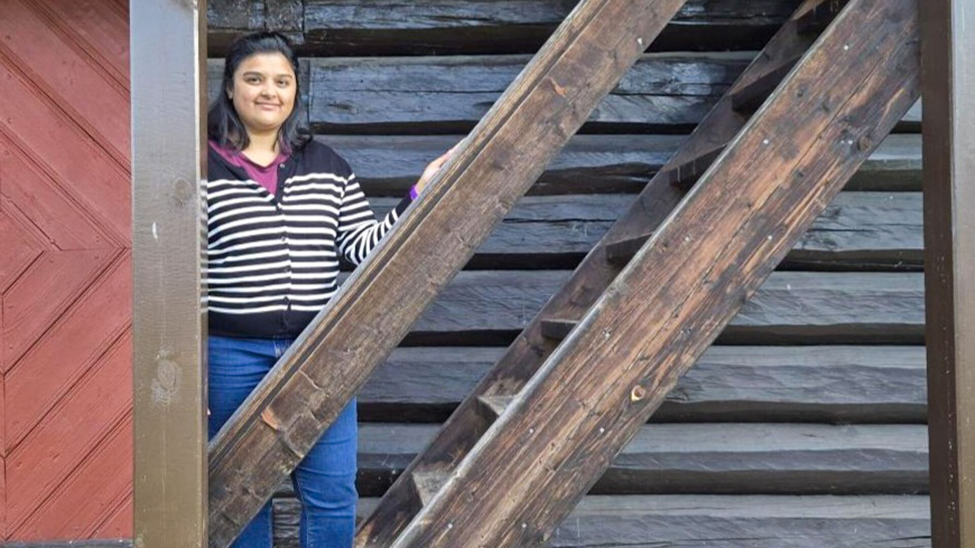 A young woman with long, black hair wearing a striped sweater and jeans standing on the stairs of an old wooden house