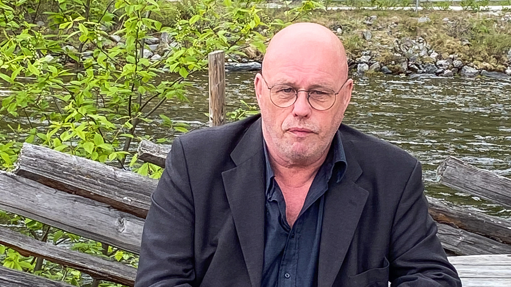 Photo of  Anders on a bench in front of a wooden fence with a river behind it.
