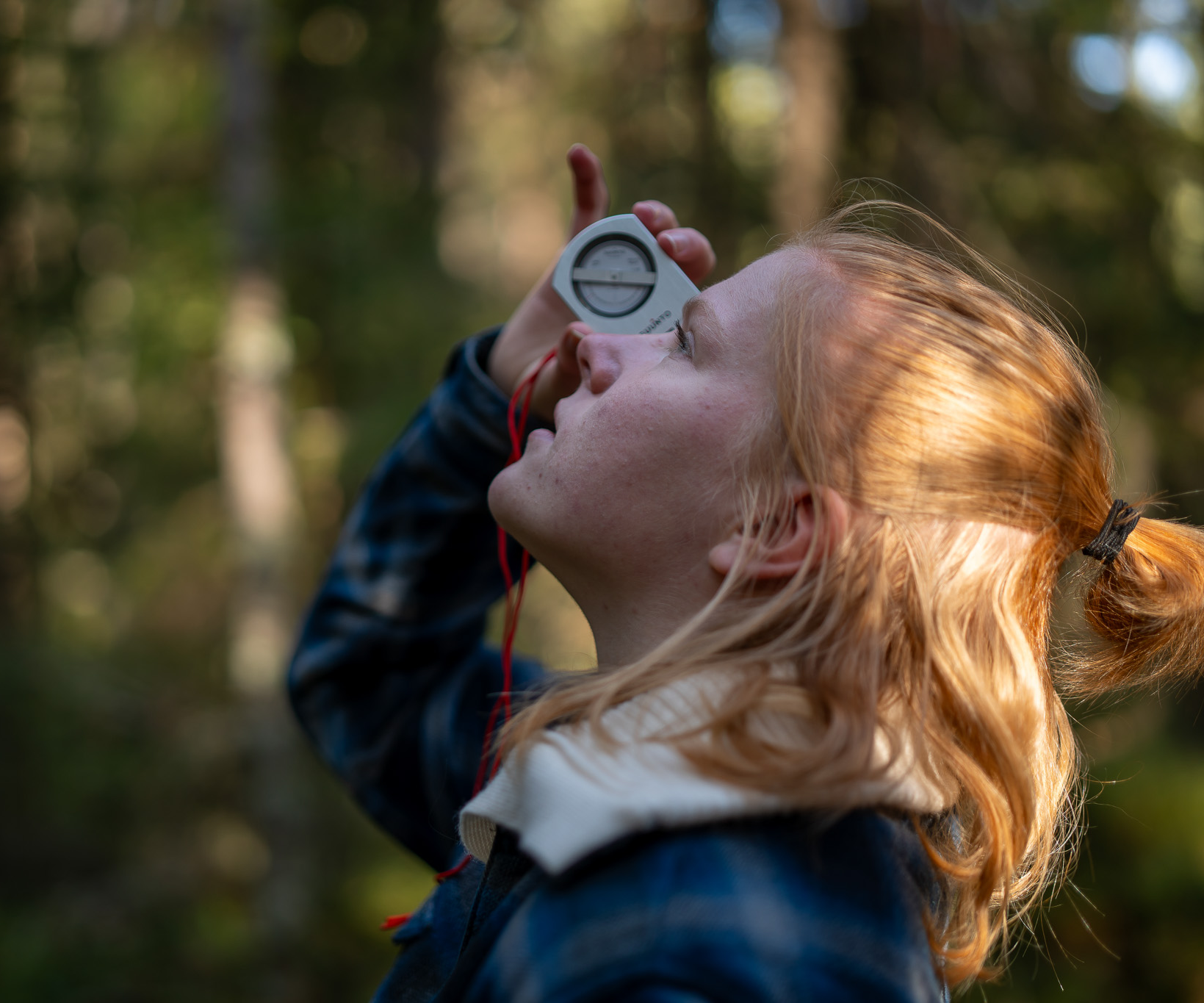 Studenten Karoline Prøis Kagnes is looking up in the air through a measuring instrument. Forest in the background