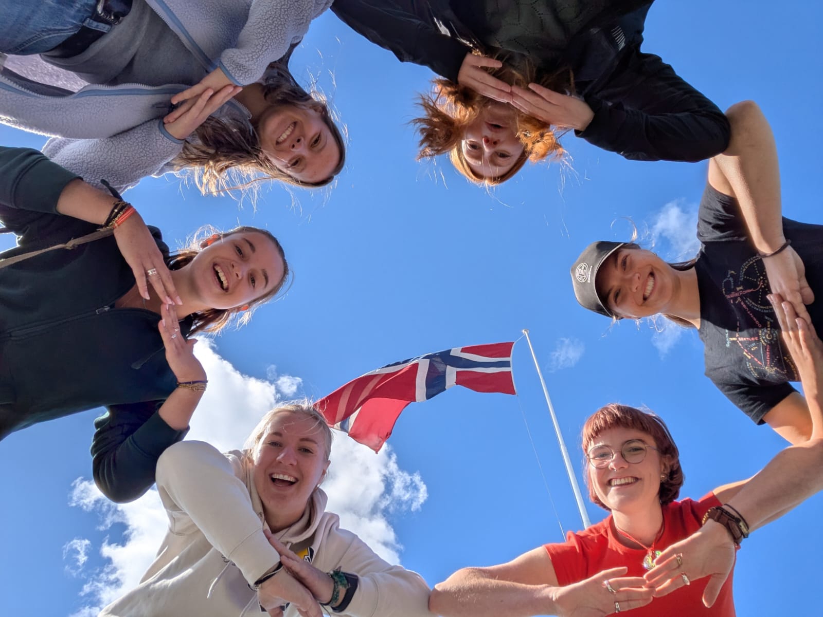 Female exchange students standing in a ring, taking a photo up towards the sky and the Norwegian flag, while posing together.