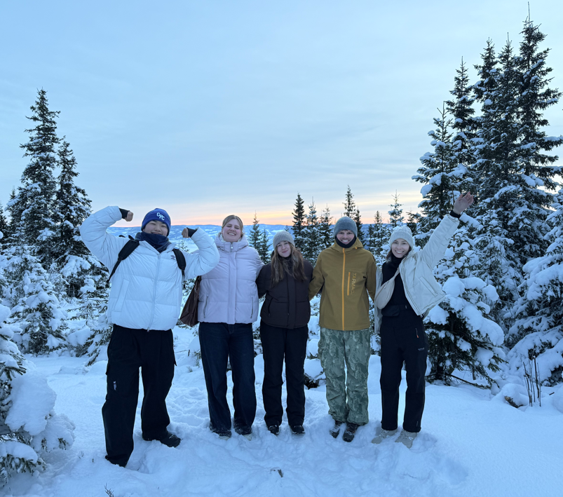 Exchange students posing happily on top of a snow covered mountains, with snow covered trees in the backround, and a yellow sunset