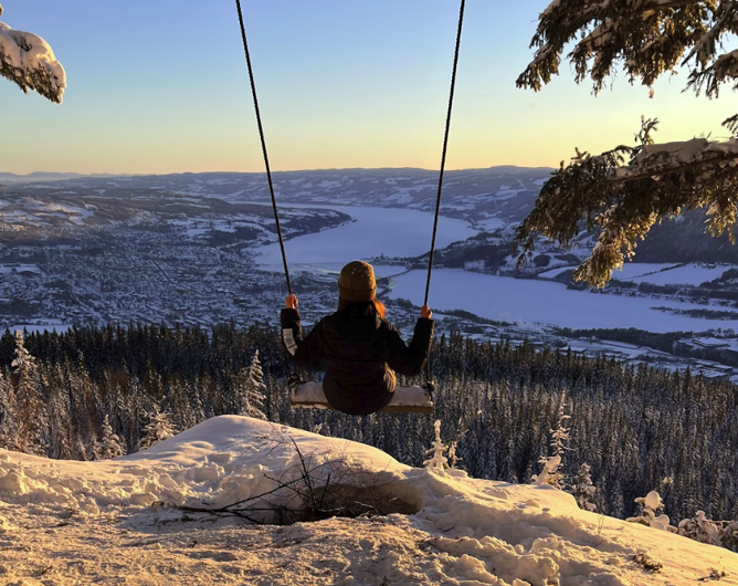 Eriko dressed in winter clothes on the swing on top of Balbergkampen during winter, with a view of Lillehammer.