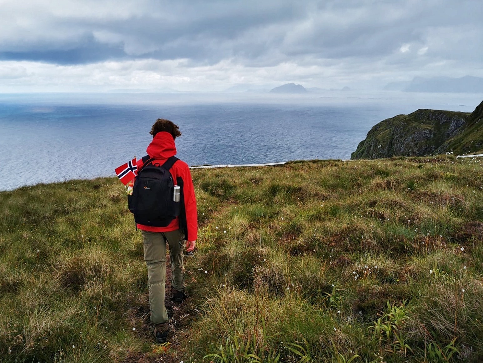 Student with the back facing the camera, overlooking the edge of a cliff towards the ocean with a Norwegian flag in the backpackl