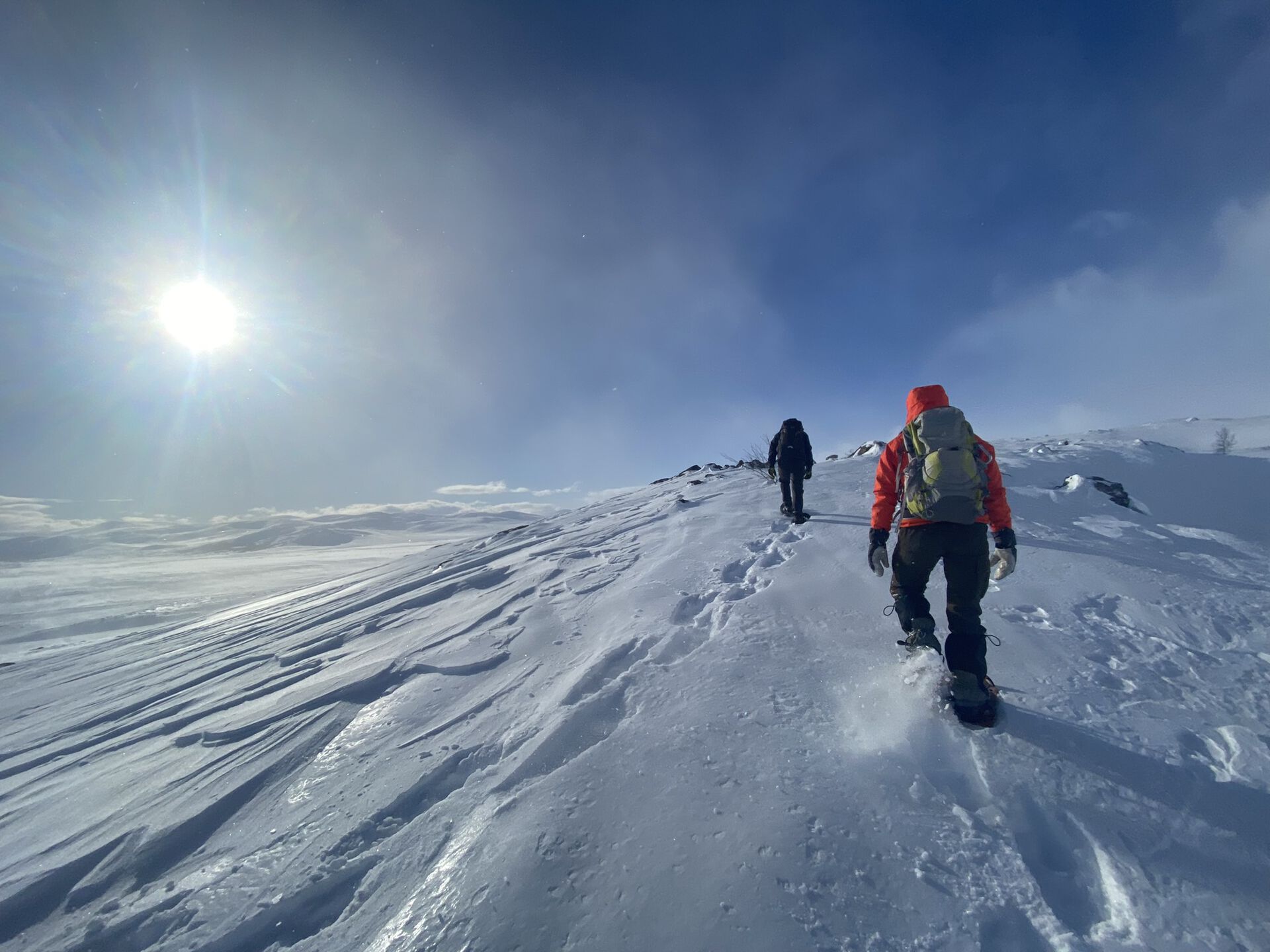Two people trudging up a snow covered mountain in snow shoes, with blue skies and sunshine