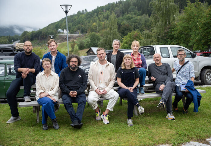 De åtte stipendiatene, phd leder og prodekan sitter poserer sammen sittende i avslappet positur på en benk ute.