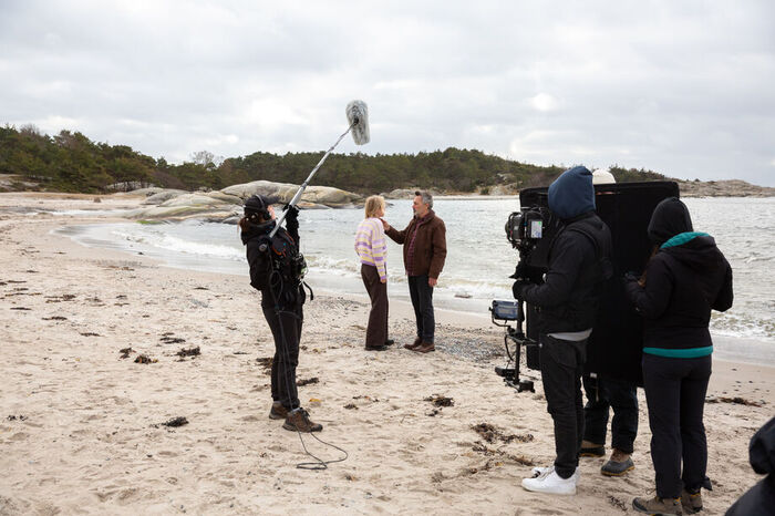 Et filmcrew filmer en mann og en ung kvinne kledd i varme klær som står og snakker med hverandre på en sandsstrand.