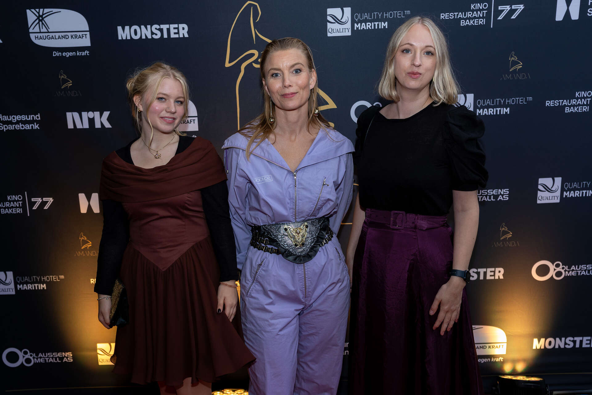 Aasne Vaa Greibrokk i lys lilla buksedress poserer på rød løper sammen med to kvinner i mørk lilla kjoler.