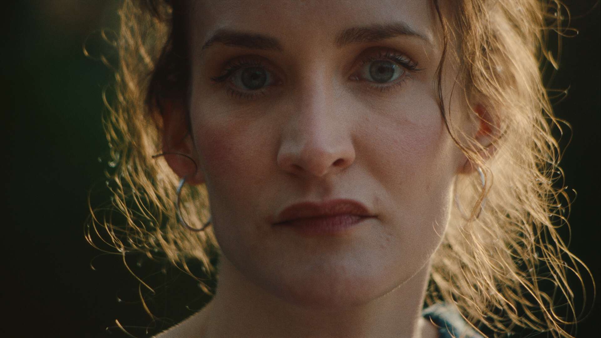 Still fra filmen "Hildring", et nærbilde av en kvinne med lett bekymret uttrykk med løst oppsatt hår opplyst av sollys bakfra.