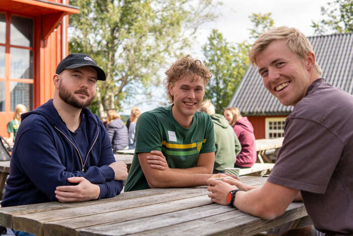 Tre smilende menn som sitter på en benk. 