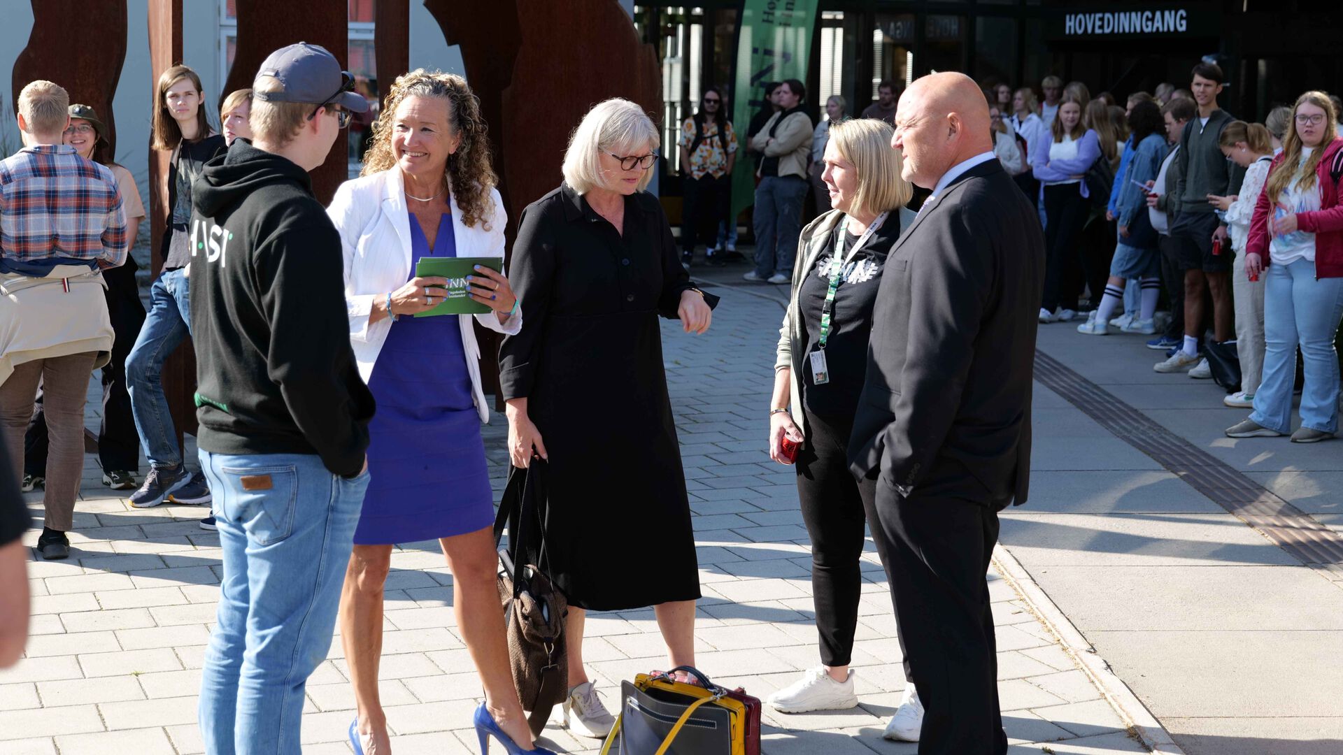 Fem personer står i gruppe og snakker, studenter står i bakgrunnen, foran hovedinngangen til studiested Hamar.