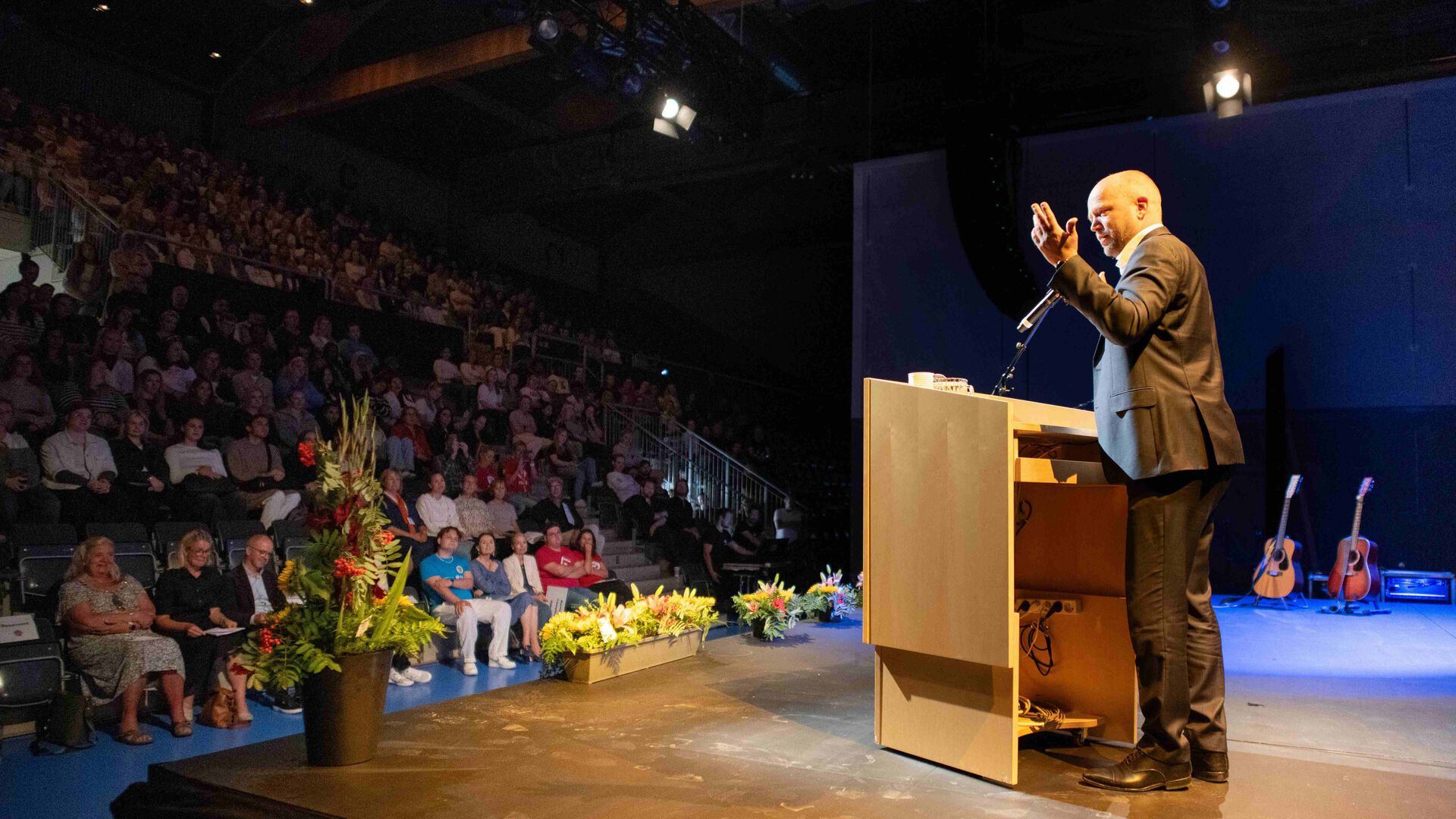 Finansministeren står på en talerstol og snakker til publikum inne i en ganske mørk idrettshall