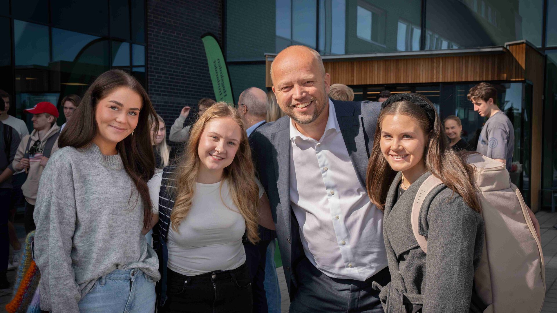 Finansministeren sammen med tre studenter, flere studenter i bakgrunnen, ute foran hovedinngangen på studiested Elverum