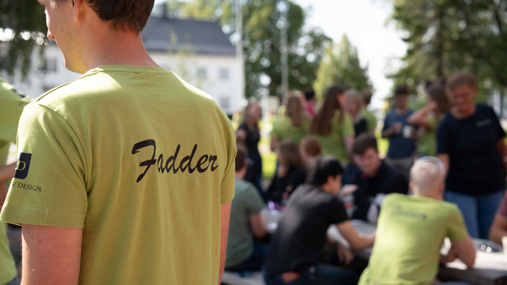 En grønn t-skjorterygg det står Fadder på, uskarpe folk i bakgrunnen