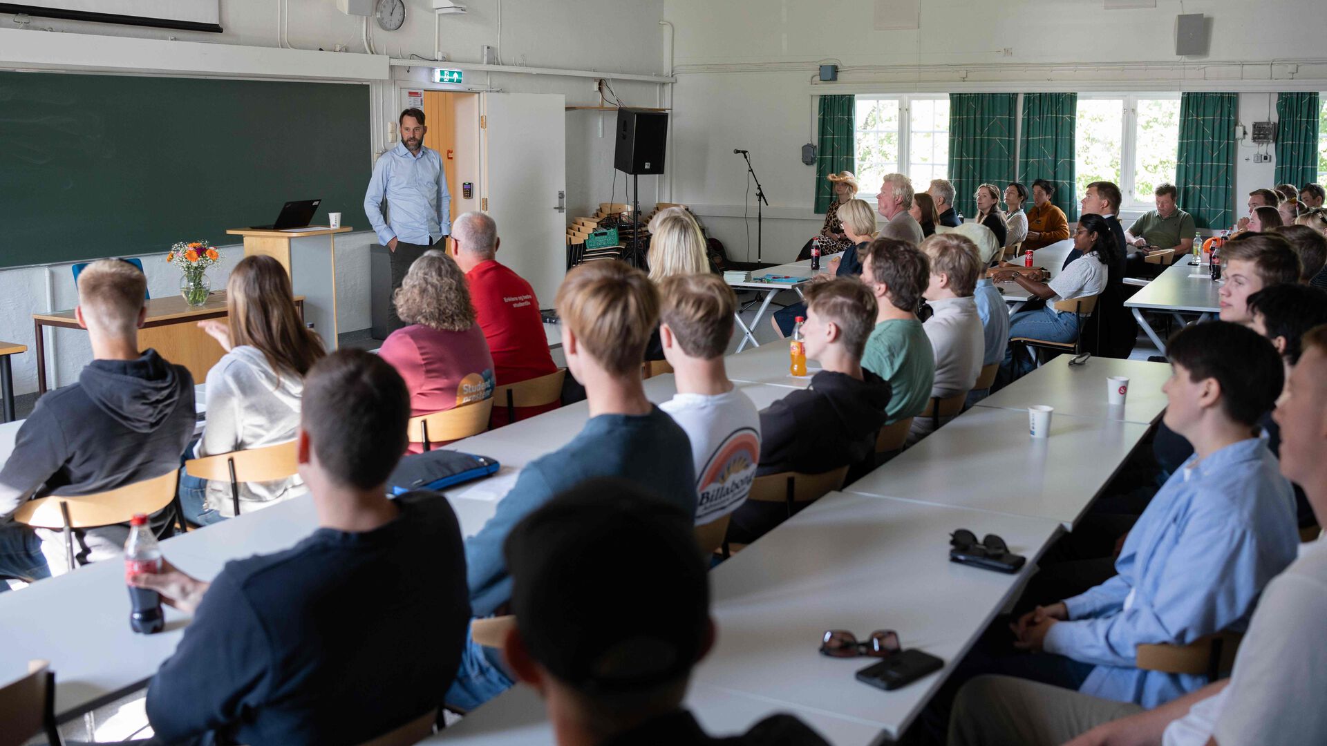 Klasserom med mange studenter på rekke og rad, en lærer foran. 