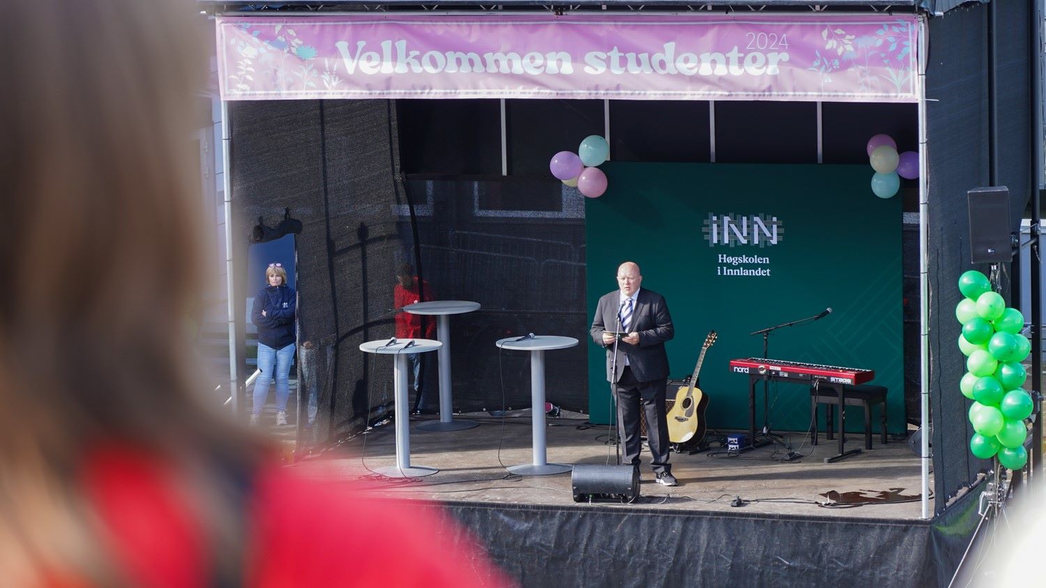 Rektor står og holder tale på en scene, folk i forgrunnen.
