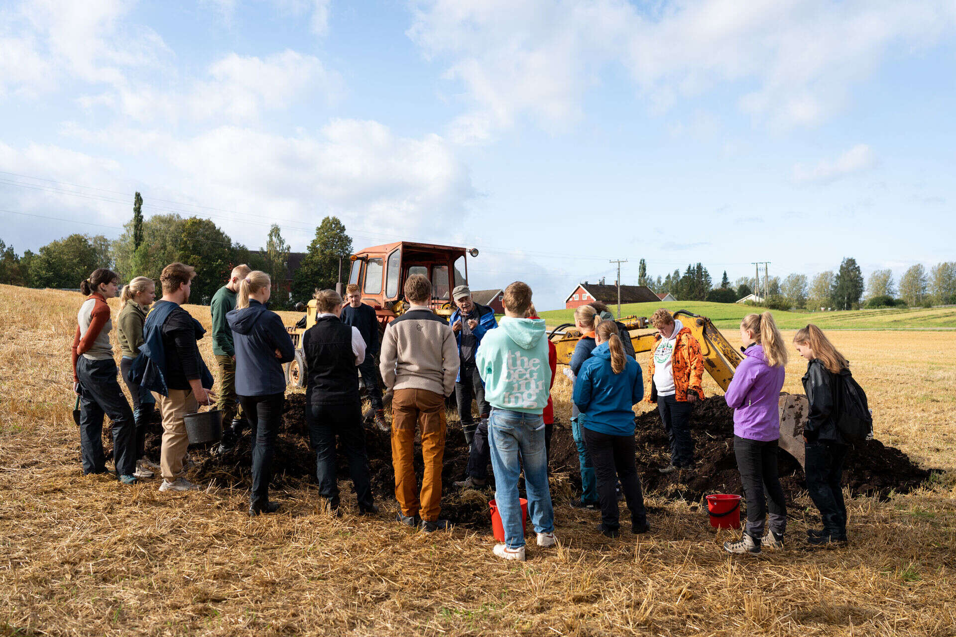 Studenter på Blæstad har uteundervisning på et jorde. De står i ring rund foreleser. En traktor i bakgrunn og blå himmel med lette skyer.