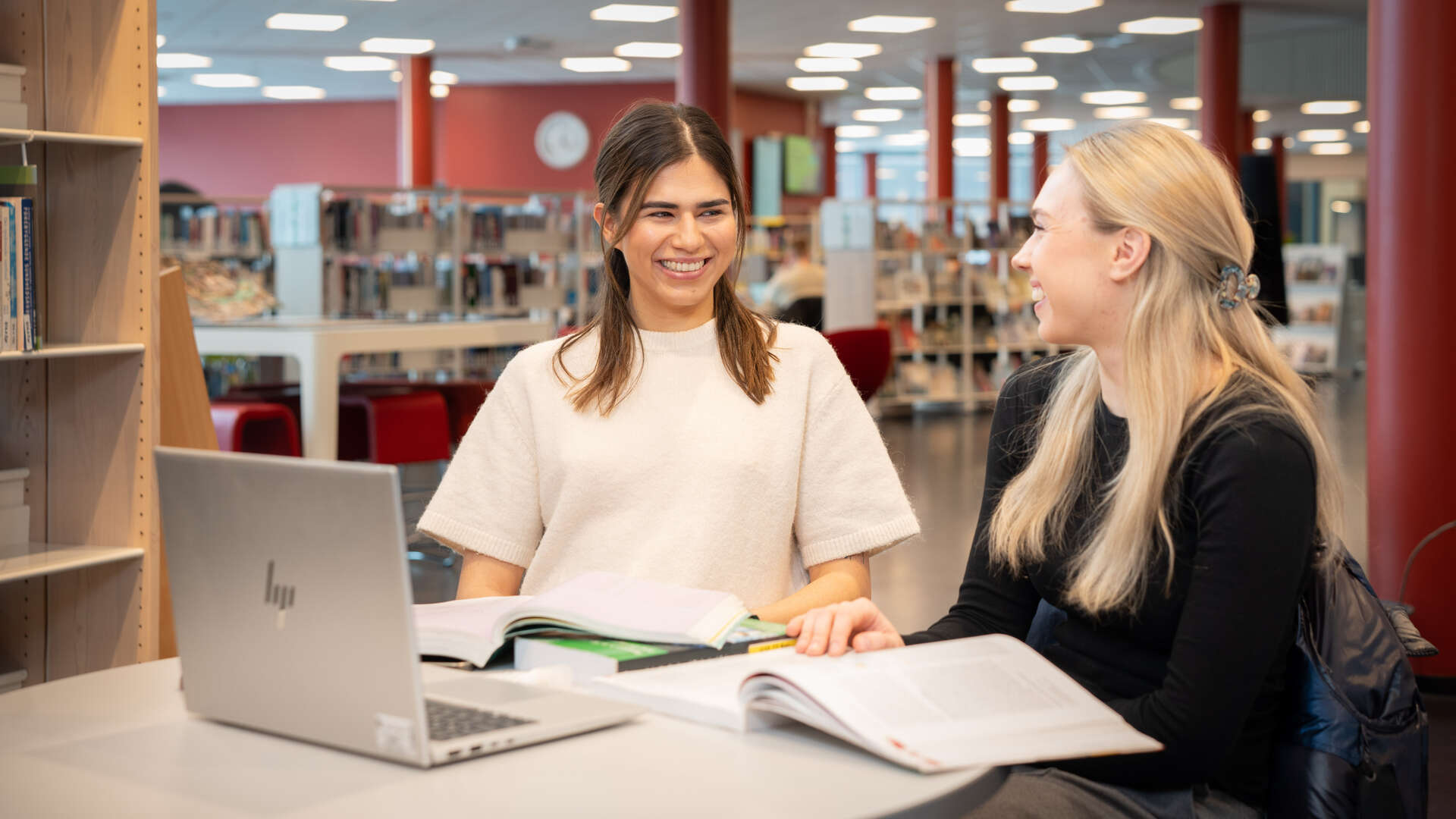 To kvinnelige studenter smiler til hverandre. De sitter ved siden av hverandre foran hver sin pc.
