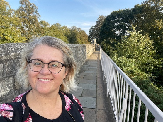 Kvinne stående foran en mur med trær i bakgrunnen.