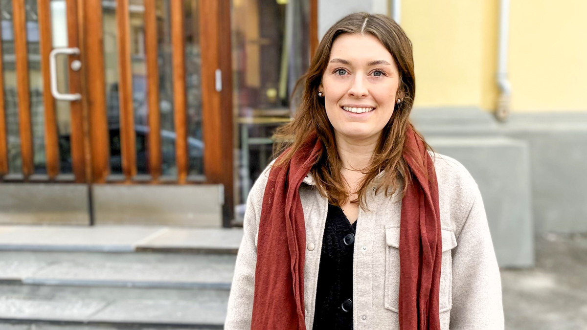Synnøve med grå jakke og rødt skjerf mot en trapp og en gul murbygning