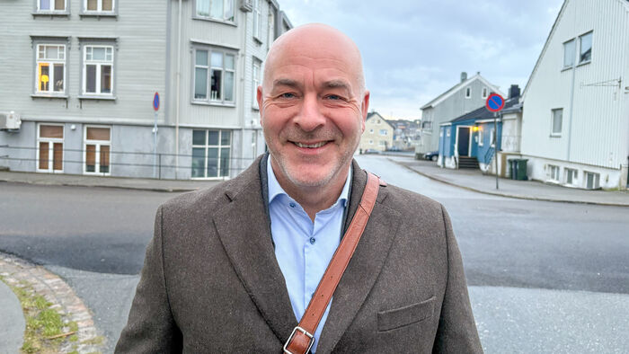 Mann med lyseblå skjorte og brun blazer og en skinnveske over skuldra står i et veikryss i en by med trehus i bakgrunnen