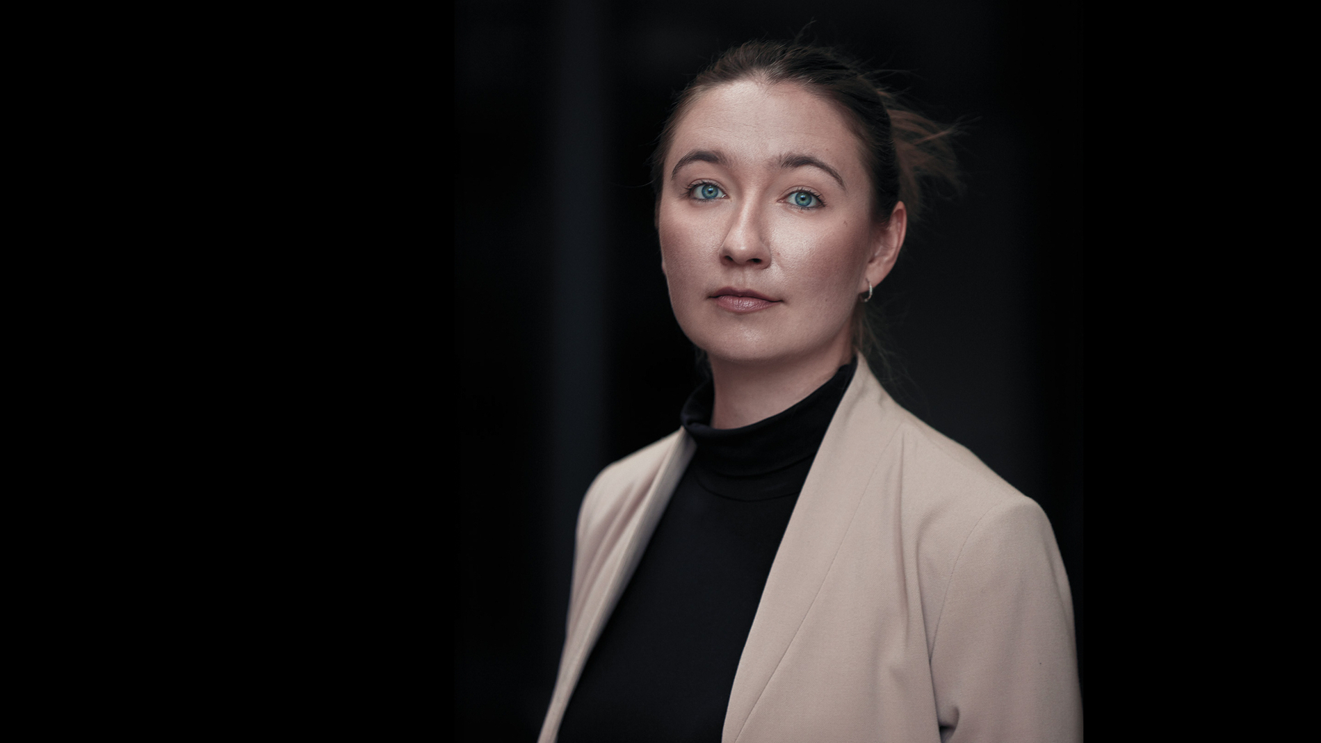 Portrettbilde av dame med svart høyhalset genser og beige blazer mot svart bakgrunn